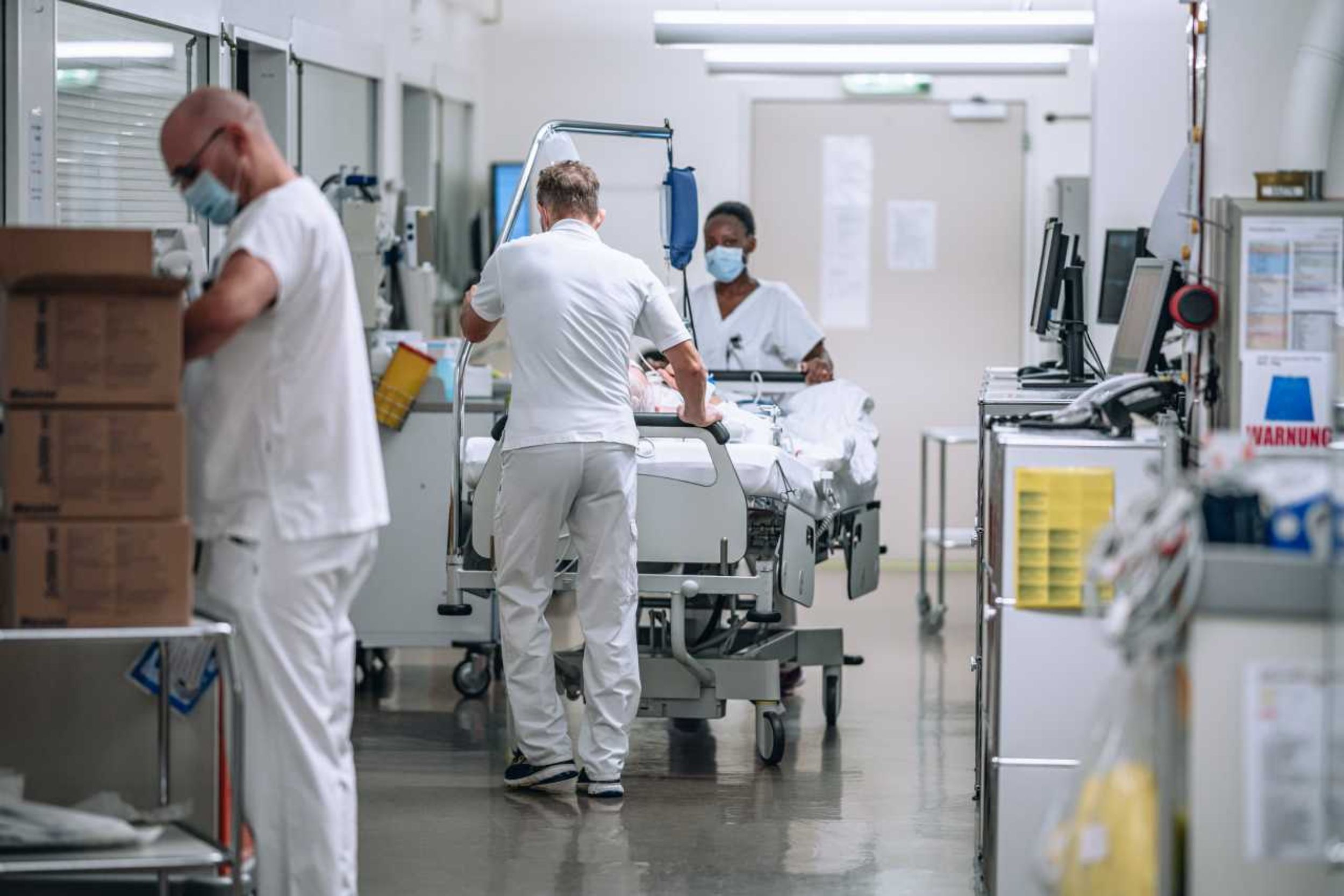 Eine Szene in einer Spitalumgebung zeigt medizinisches Personal in weisser Berufskleidung, das sich um einen Patienten auf einem Bett kümmert. Ein Mann schiebt das Bett durch einen Korridor, während eine weitere Person mit einer Maske das Bett begleitet. Im Vordergrund steht ein weiterer Mitarbeiter an einem Tisch. Die Umgebung ist klar strukturiert, mit medizinischen Geräten, Monitoren und Aufbewahrungssystemen. Die Szene zeigt eine Intensiv- bzw. Notfallstation.
