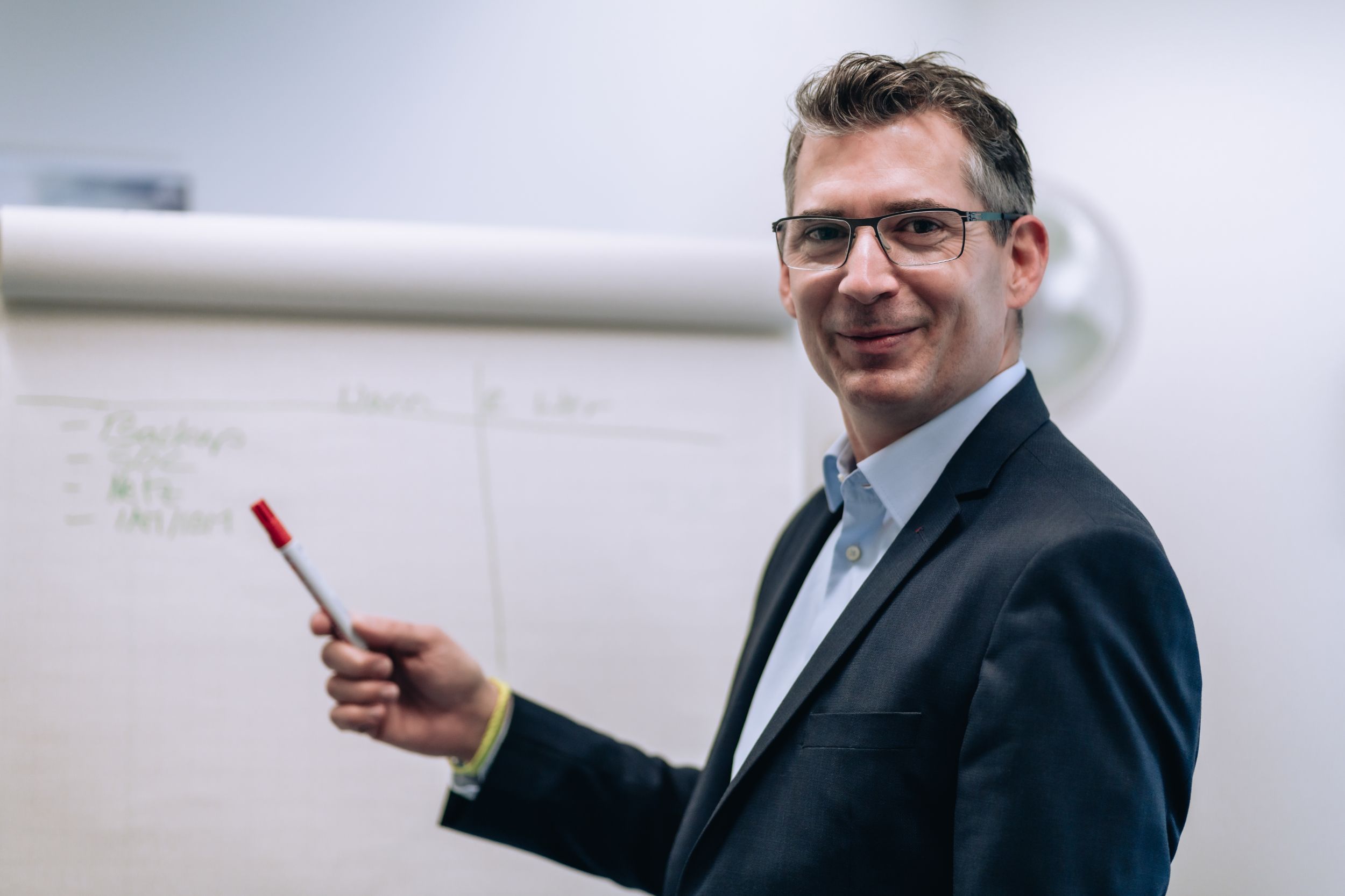 Mann mit Brille und Anzug zeigt mit einem Stift auf ein Flipchart