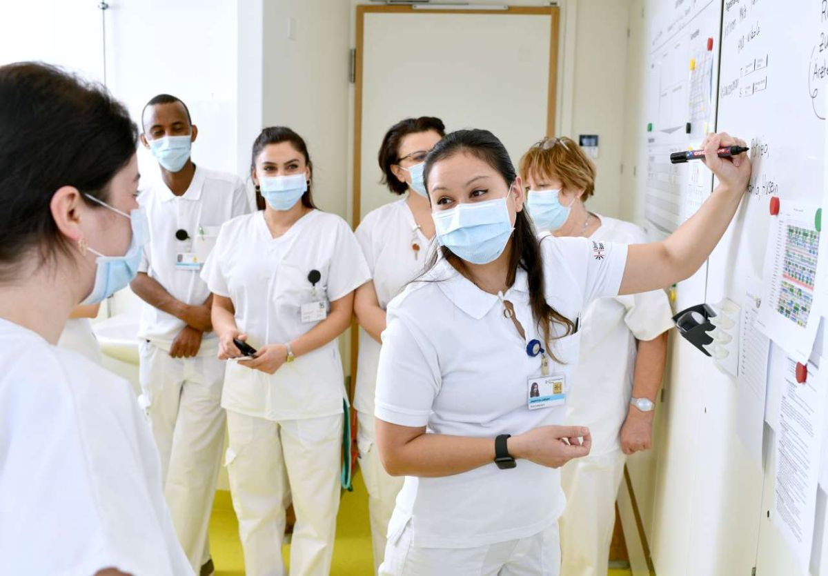 Pflegefachpersonen in weissen Uniformen und mit Gesichtsmasken stehen in einer Gruppe vor einem Whiteboard in einem Spital. Eine Frau zeigt mit einem Stift auf das Board und erklärt etwas, während die anderen aufmerksam zuhören. Die Szene wirkt professionell und konzentriert, typischerweise im Rahmen einer Team- oder Übergabebesprechung.