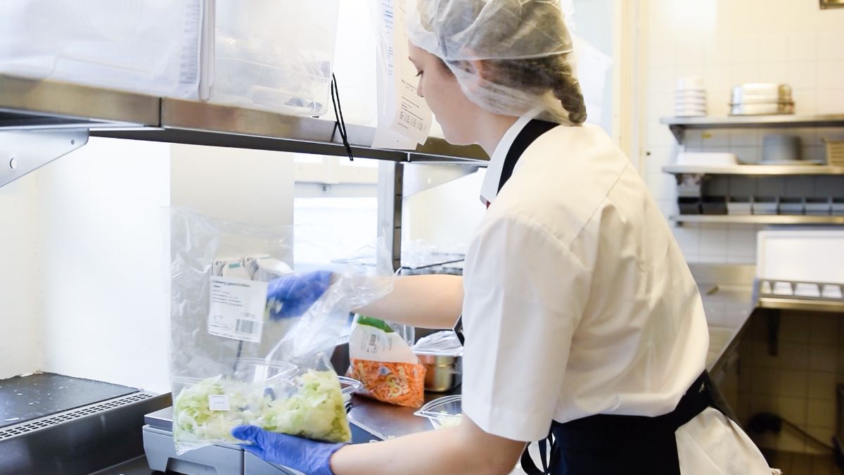 Eine Fachfrau Systemgastronomie in Ausbildung steht lächelnd hinter der Theke und serviert einen Kuchen